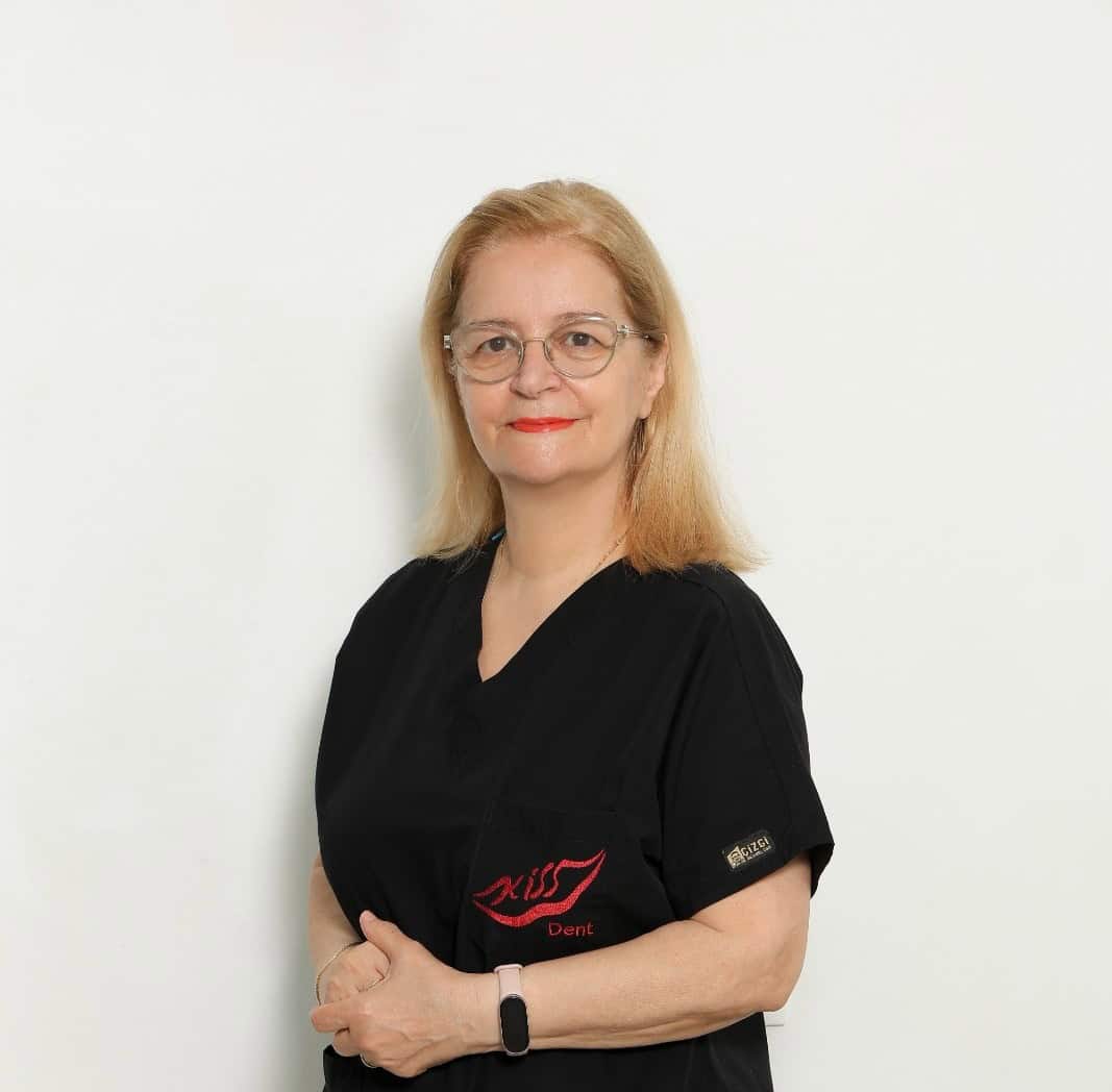 Dentiste femër me uniformë të zezë me syze duke buzëqeshur për kamerën.